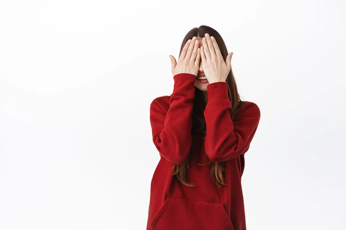 cute caucasian woman red hoodie covering her face eyes with palms smiling waiting surprise playing peekaboo hide n seek standing against white wall